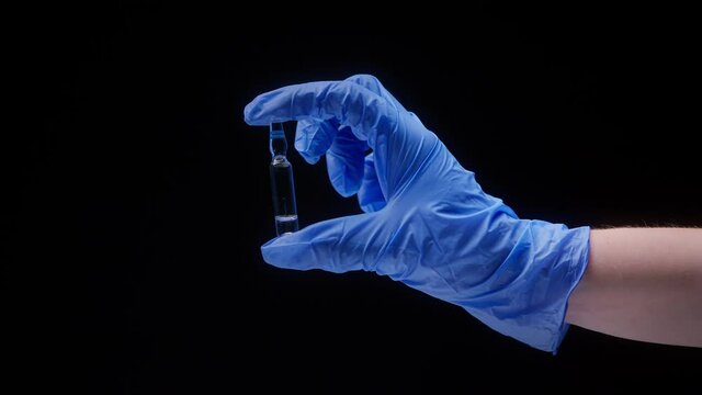 Close-up of hand wearing blue medical glove, holding ampoule with vaccine against coronavirus. Nurse showing sample of medicine on black background, vaccination against covid, laboratory research. 