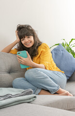 Obraz premium Young latin brunette smiling, casually dressed with yellow blouse and blue jean, looking at her smartphone while resting his head on one hand, sitting on the sofa