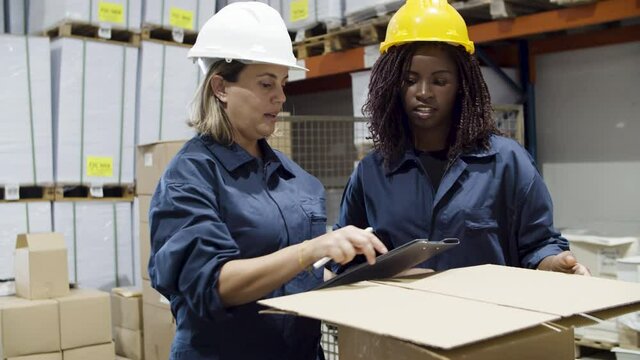 Female workers of logistic company talking in warehouse. Caucasian woman holding clipboard, explaining her African American coworker process of distributing goods. Manufacture, factory concept.