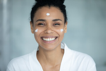 Happy overjoyed mixed race woman with moisturizing collagen cream spots on smooth healthy skin looking at camera with toothy smile, enjoying home skincare treatment and spa. Head shot portrait