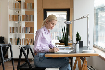 Side view confident businesswoman taking notes, writing in notebook, sitting at desk with laptop, employee intern watching webinar, working on research project in modern office, planning workday