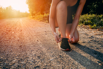 young women in sportswear sitting and tying shoelaces her shoes before start running on gravel at sunset way in the park, female training to a trail run or marathon outdoor, healthy wellness concept.