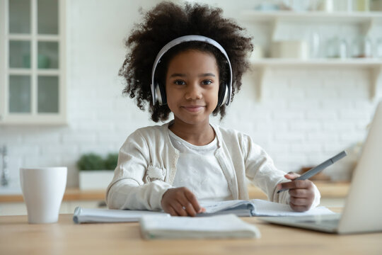 Portrait of smart small 7s African American schoolgirl in earphones study distant on computer at home. Happy little biracial girl child in headphones have online webcam virtual lesson on laptop.