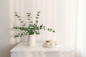 Bouquet of eucalyptus branches in vase and tray with candles standing on the chest of drawers in the morning sun beams. Minimalistic home decor. White stylish interior.