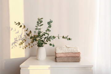Bouquet of eucalyptus branches in vase and stack of knitted sweaters in pastel colors standing on the chest of drawers in the morning sun beams. Minimalistic home decor. White stylish interior.