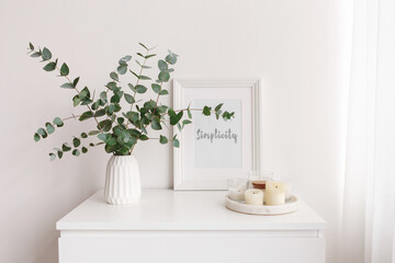 Frame with text SIMPLICITY, bouquet of eucalyptus branches in vase and tray with candles standing on the chest of drawers. Minimalistic home decor. White stylish interior.