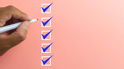 Close up Left hand holding blue pen with check box on pink pastel background with copyspace.,checklist concept.