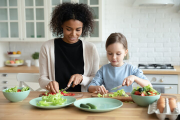 Happy loving African American mother and little Caucasian adopted daughter have fun prepare healthy...
