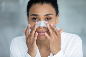 Portrait of positive beautiful Black woman during skincare cleansing procedures. Girl applying sticky strip on nose for peeling skin, cleaning pores, removing blackheads, prevention acne. Close up