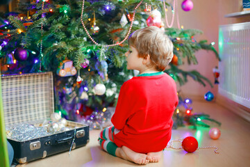 Fototapeta na wymiar Little child decorating the Christmas tree with balls. Cute happy preschool boy in nightwear pajamas sitting by tree with traditional decorations. Xmas Eve concept. Kid with lights on background
