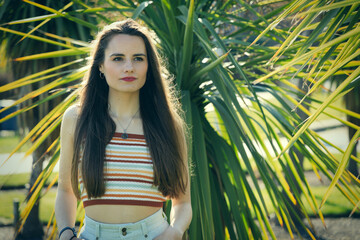 Portrait of a young brunette woman standing near a palm tree in a park on a sunny day
