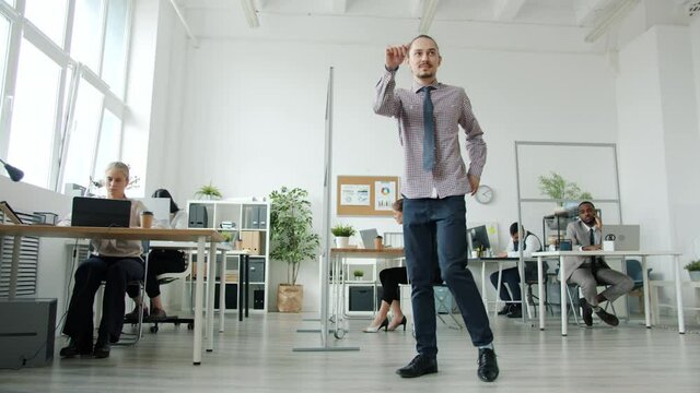 Slow motion portrait of funny hipster man dancing at work with serious people sitting at desks in background. Performance and business activities concept.