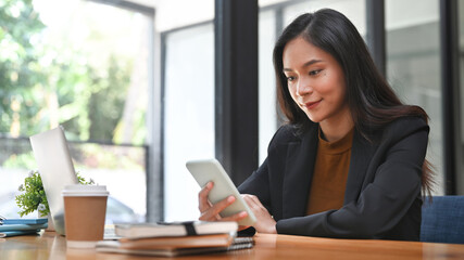 Happy businesswoman sitting in modern workplace and using mobile phone.