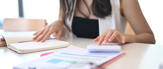Cropped shot of female accountant analyzing finance data and using calculator on white office desk.