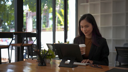 Fototapeta premium Happy businesswoman drinking coffee and reading news on computer tablet at office in the morning.