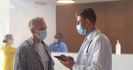 Male doctor and mature man wearing protective face masks communicating in waiting room at hospital.