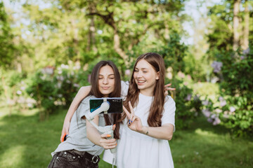 Smiling pretty young female influencer bloggers are filming or recording video with their mobile phone on a stabilizer, in a sunny green park outside. Blogging concept. Soft selective focus, defocus.