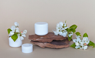 White jars of skincare cosmetics with fruit extract on a stone plinth and apple blossoms nearby. Cosmetics with apple extract. Spring skin care for face and hands. Front view, close-up, copy space