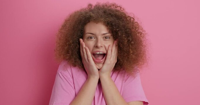 Surprised cheerful European woman keeps hands on cheeks being impressed by something unbelivable smiles broadly dressed casually poses against pink background. Happy human reactions concept.