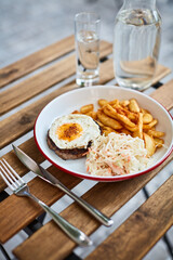 schnitzel with egg, potatoes and cabbage on a wooden table on the street terrace summer with a fork and knife with a glass jug of water