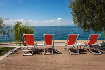 Four empty deck chairs on a beautiful beach on Lake Garda (Lago di Garda) between the small village of Bardolino and Garda town. Verona province, Veneto, Italy, Europe. Lombardy coast on the horizon.
