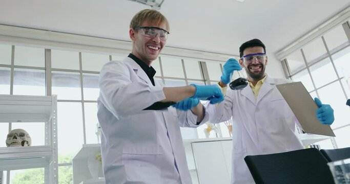 two young funny male scientists wearing white lab coat dance around in the science lab holding glass flask with chemical inside. Concept of crazy mad scientist lab experimentation.