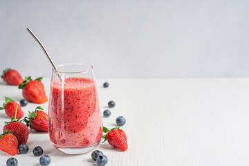 Refreshing organic smoothie drink made from pureed ripe sweet juicy strawberries and blueberries served in tall drinking glass with metal reusable straw  on white wooden table. Image with copy space