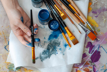 The artist's close-up hand holds a brush against the background of a palette, brushes and paints, is engaged in art, creativity, hobbies, anti-stress or art therapy.