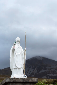 White statue of Saint Patrick holding green shamrock at the start of foot path to the peak of Croagh Patrick, county Mayo, Ireland. Mountain peak in the background