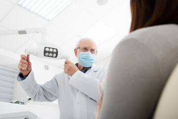 senior dentist in medical mask holding medical lamp near patient sitting in dental chair.