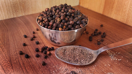 Black pepper in a bowl along with powdered pepper in a spoon in wooden background