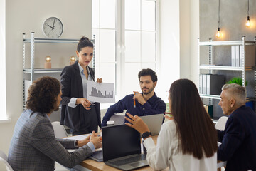 Strict team manager standing at office table in corporate group meeting, holding column chart paper, showing sales increase decrease figures and listening to female coworker making smart suggestion