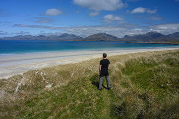 Man gazing out over white sands and turquoise water of Luskentyre beach in Isle of Harris, Outer Hebrides, Scotland. Mountains in background.