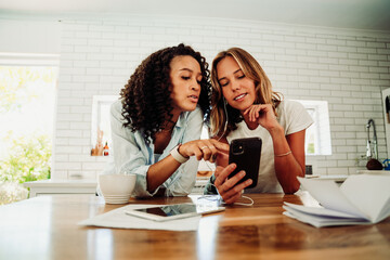 Two mixed race friends relaxing in kitchen looking at cellular device while working from home