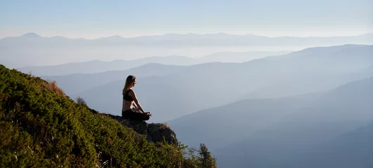 Poster Im Rahmen Fit young woman sitting on grassy hill in lotus position and doing Gyan mudra hand gesture while mediating outdoors. Sporty woman doing meditation yoga exercise in mountains. Copy space. © anatoliy_gleb