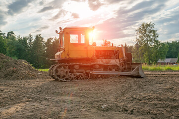 An old yellow rusty excavator is clearing a plot of land. The excavator works at sunset
