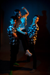 Two guys in a plaid shirt are holding a girl on a dark background in a studio with multicolored light