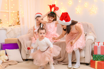 children girls portrait, sisters dressed in santa helper hat, sitting on a couch in home interior decorated with christmas lights and new year holiday gifts