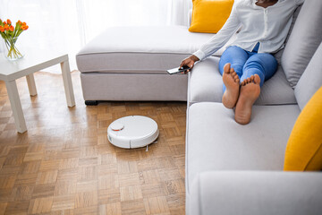 Robotic vacuum cleaner cleaning the room while woman relaxing on sofa. Woman controlling vacuum with smart phone. Woman relaxing on sofa while robot vacuum cleaner doing housework