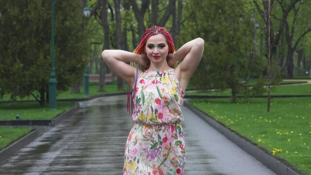 A girl with a happy smile in a dress with African braids and make-up whirls and throws her hair up in spring rainy park