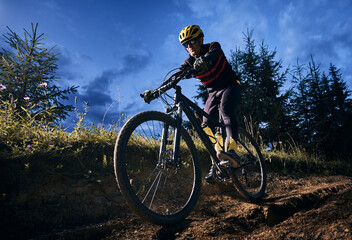 Wide angle view of young man riding bicycle downhill with blue evening sky on background. Bicyclist...