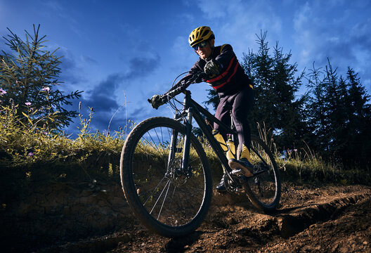 Wide angle view of young man riding bicycle downhill with blue evening sky on background. Bicyclist in cycling suit cycling down grassy hill at night. Concept of sport, biking and active leisure.