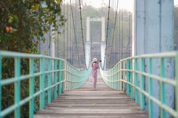 A female tourist in a pink dress holds a camera walking on a suspension bridge, a wooden floor with a light green iron handle. Located in Thailand