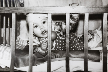 Little baby in bed looking through the wooden rods. Vintage black and white paper photo. Early...