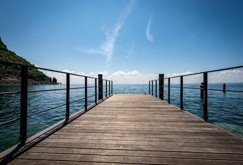 Fototapeta premium Small empty wooden pier on Lake Garda (Lago di Garda) in front of the small town of Garda, tourist resort in Verona province, Veneto, Italy, southern Europe.