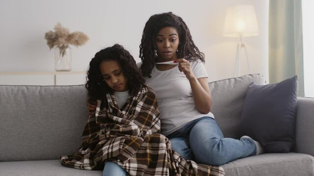 Worried caring african american mother checking temperature of her sick daughter, seeing fever and embracing kid