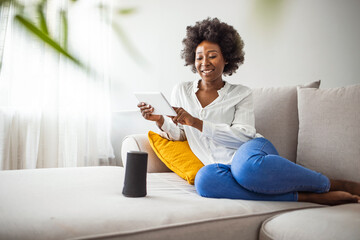 Happy young woman controlling smart home devices with a voice commands and smart speaker at home. Concept of a smart home and managing wireless devices remotely