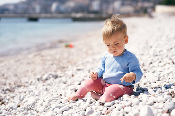 Pensive kid sits on a pebble beach, looking at his feet, near the sea
