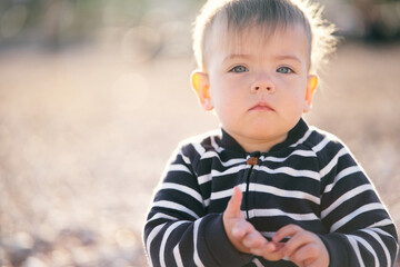 Serious toddler in striped overalls sits on a pebble beach, holding his hands in front of him. Close-up