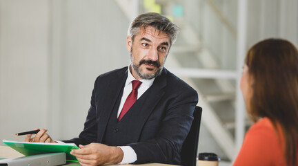 Senior Caucasian gentel businessman in black suit and red necktie sitting in meeting as boss and kindly discuss with colleague on left for asking information, question, and opinion